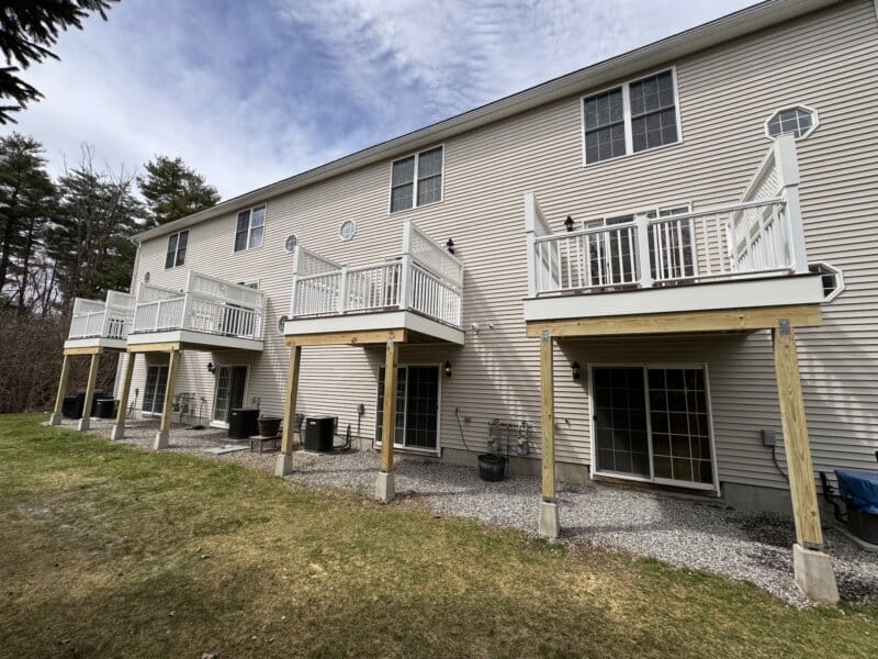 Row of three-story townhomes with freshly built elevated decks—white PVC railings and privacy panels—supported by new pressure-treated posts on concrete footings above gravel patios with sliding glass doors, beige vinyl siding, and a grassy common yard under a partly cloudy sky.