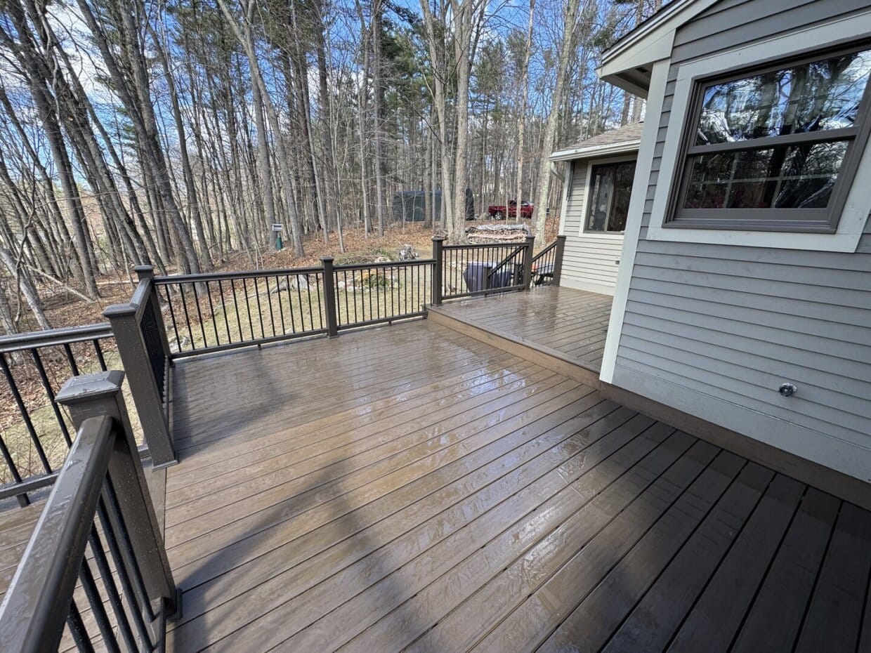 TimberTech composite deck with black railings attached to a gray house, overlooking a wooded backyard with tall trees and patches of grass.