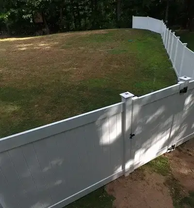White vinyl privacy fence enclosing a sloped backyard, with a secure gate featuring a black latch and hinges, creating a clean and durable boundary against the wooded backdrop.