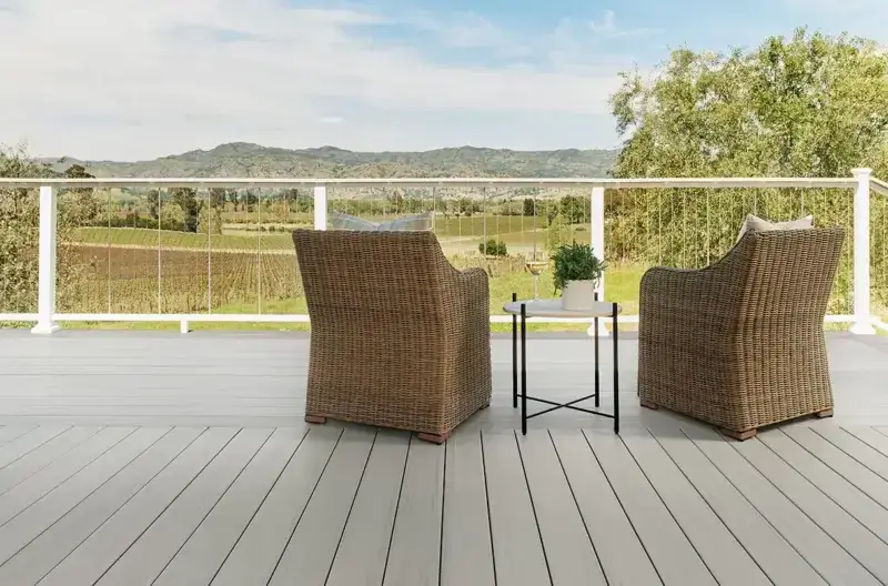 Light gray TimberTech composite deck with white cable railing, two wicker armchairs, and a small round side table with plant and wine glass; open fields and hills beyond.