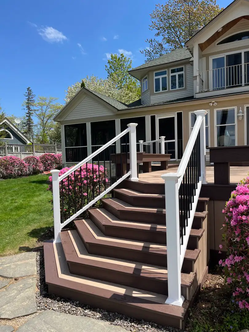 Wide brown composite deck stairs with angled treads, white posts and rails with black balusters, flanked by blooming pink shrubs; large house and green lawn under blue sky.