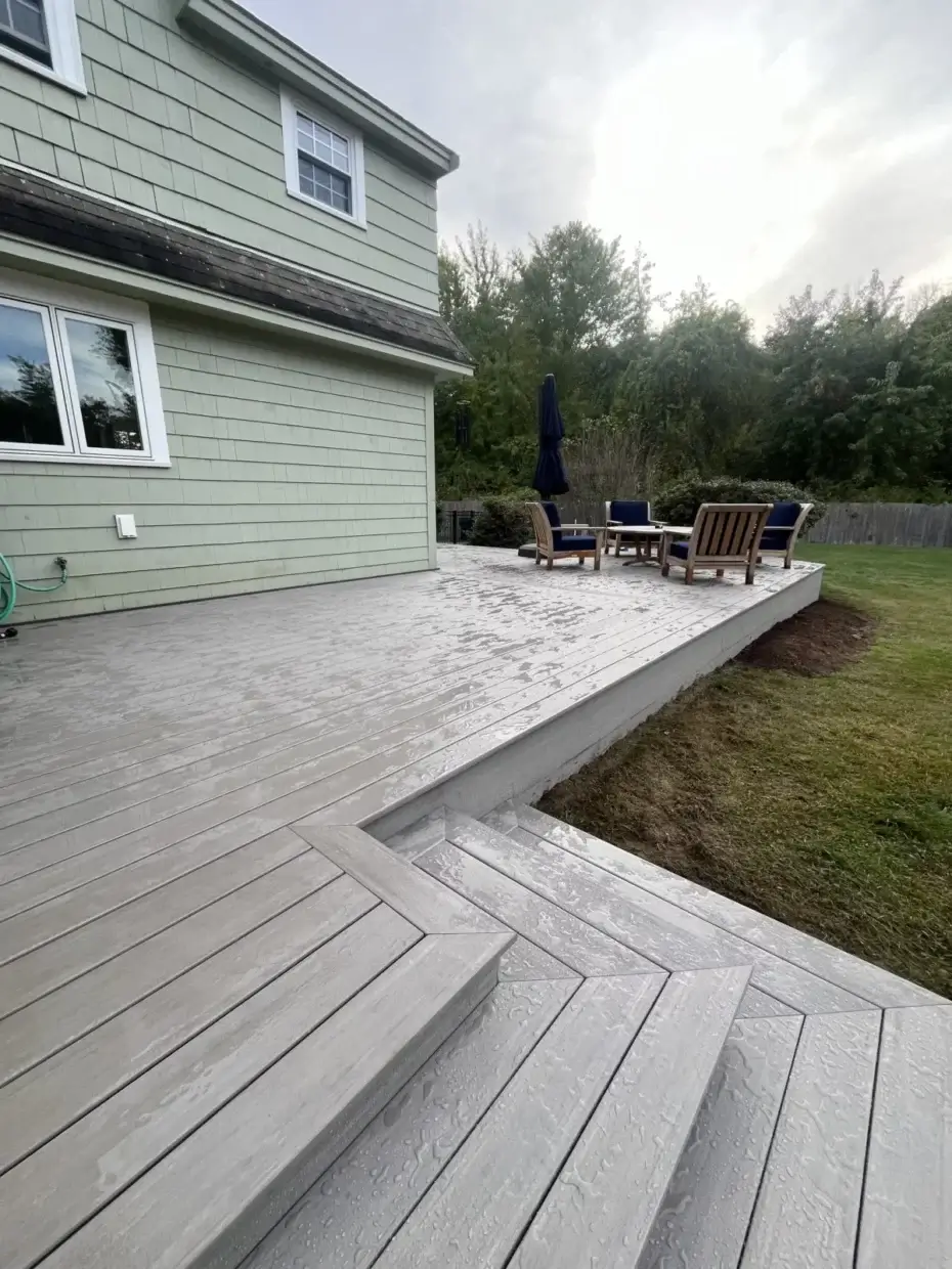 A light gray composite deck with wide steps leading down to a lower seating area, featuring outdoor chairs and an umbrella beside a green shingle-sided home with a grassy backyard.