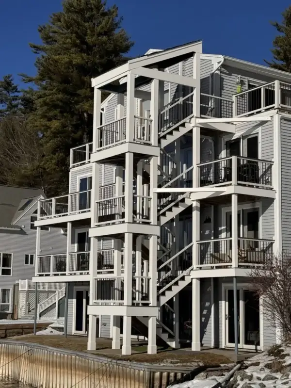 Multi-story waterfront home featuring stacked white decks with black railings and exterior staircases, overlooking a shoreline with snow patches and tall pine trees under a clear blue sky.