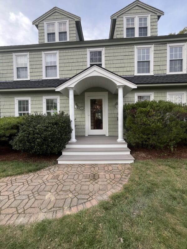 A classic front entry with a small composite porch, white columns, and steps leading to a centered front door, set against a green shingle-style home with dormer windows and landscaped shrubs.