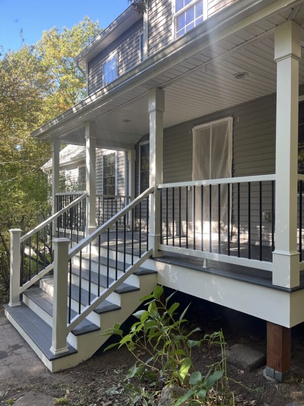 A covered front porch with composite decking, white square columns, black metal balusters, and a short staircase, attached to a gray home and surrounded by trees and landscaping.