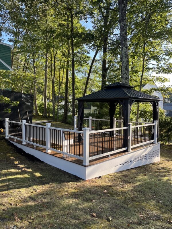 A freestanding composite deck with white PVC-wrapped framing, black balusters, a built-in bench, and a black screened gazebo, set in a wooded backyard with mature trees and surrounding lawn.
