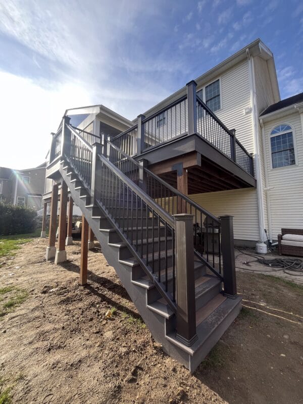 A newly constructed elevated composite deck with gray decking, black metal balusters, wide staircase, and privacy screen, built alongside a light-colored home with reinforced posts and clean modern railings.